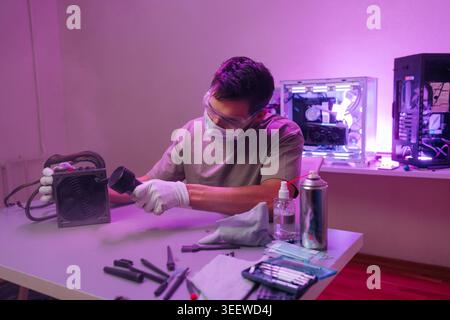 Young man wearing protective mask, gloves glasses performing maintenance on computer power supply, blowing dust away with compressed air at workspace Stock Photo