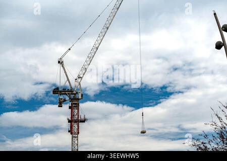 Tower Crane Lifting Load Against Sky, Metal Lattice Boom Rising Above Urban Construction Site With Cloudy Blue Backdrop, Distant Buildings Stock Photo