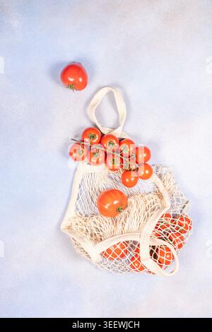 Ripe red tomatoes, including cherry tomatoes on the vine, sitting in and around a reusable mesh shopping bag on a light blue background, promoting sus Stock Photo