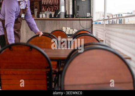 Empty Wooden Tables And Stacked Chairs, Waiter In Purple Shirt And Apron Arranging Seating On Rooftop Terrace During Closing Shift, Soft Evening Stock Photo