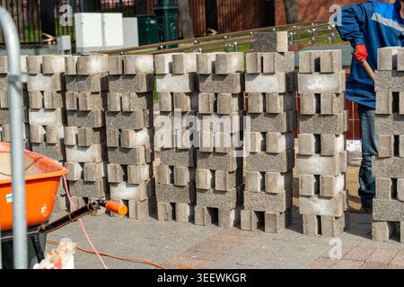 Stacked Concrete Paving Blocks On Site, Worker Arranging Materials Beside Orange Wheelbarrow, Industrial Yard With Pallet And Equipment, Textured Gray Stock Photo