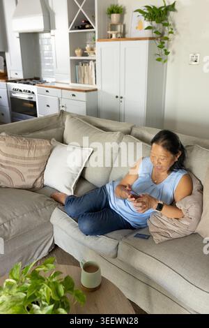 Senior Asian woman reclining on sofa, scrolling smartphone, wearing blue top, jeans in open kitchen Stock Photo