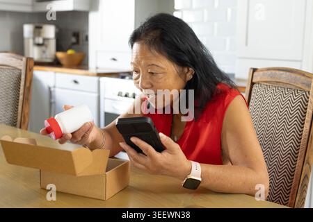 Senior Asian woman in red tank top holding red-capped bottle while checking phone at kitchen table Stock Photo