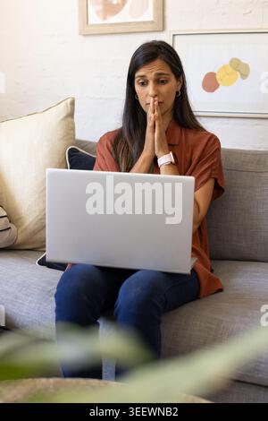 Adult Indian female sitting at home on grey sofa wearing rust shirt and jeans, using laptop Stock Photo