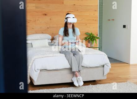 Woman wearing white VR headset sitting on edge of bed holding controller in bedroom Stock Photo