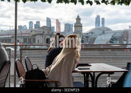 Two Women At Riverside Cafe Sharing Quiet Conversation While Overlooking City Skyline, Metal Railing In Foreground, Wicker Chairs And Small Round Stock Photo