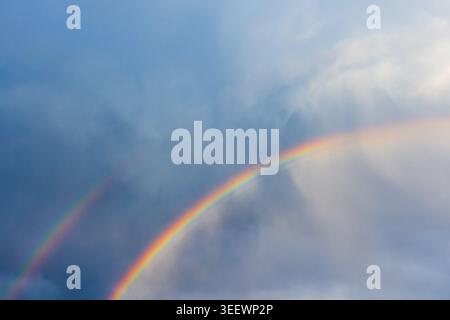 A vibrant double rainbow arcs across a moody blue sky with soft clouds and diffused light, creating a serene, hopeful atmosphere that highlights color Stock Photo