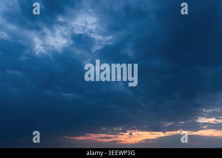 Dramatic blue storm clouds sweep across the evening sky above a thin orange sunset horizon, creating a moody, atmospheric scene ideal for weather, nat Stock Photo