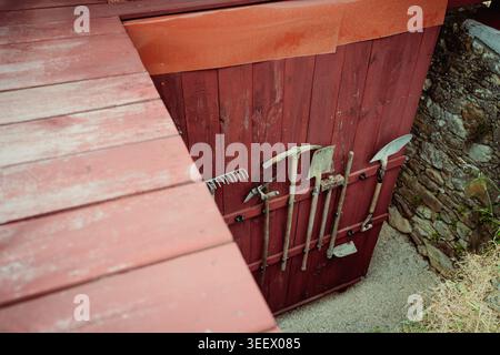 High angle rustic detail of red wooden boards, stone wall, blurred walkway, and hanging garden tools, ideal for rural workshop, storage, and texture t Stock Photo