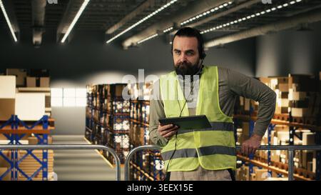 Warehouse operator, assisting clients, order fulfillment, package tracking, wearing headset and safety vest. Staff checking parcel status via airway bills on tracking software, supply chain. Stock Photo