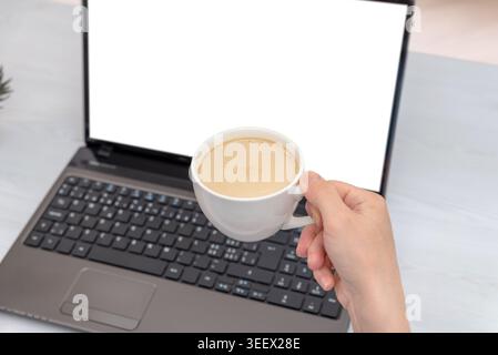 Hand holding coffee cup in front of laptop mockup screen, creating cozy remote work atmosphere with minimal office desk composition and copy space Stock Photo