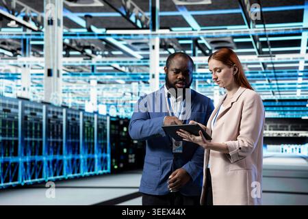 Data center engineering team working together with laptop, doing gear upgrades. Server farm teamworking colleagues using notebook to oversee operations and resource allocation Stock Photo