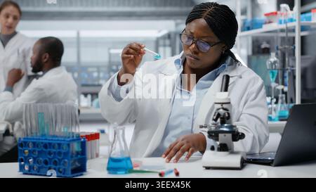 Clinical laboratory technologist pipetting reactant into blue solution for purity analysis. African american woman adding liquid agent to test tube to observe chemical reaction properties, camera B Stock Photo