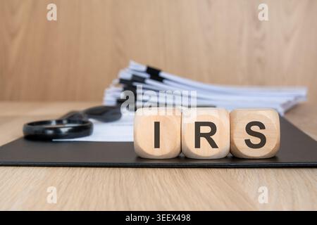 Wooden cubes with the word IRS on a black clipboard. Blurred stack of tax documents and a magnifying glass in the background. Concept for Internal Rev Stock Photo