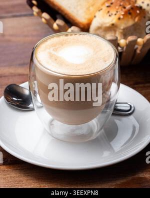 Cup of coffee with creamy foam sits on white saucer. Spoon rests beside, inviting a sip. Basket of bread looms softly in background. Warm wooden table Stock Photo