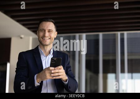 Smiling businessman using smartphone in modern office corridor Stock Photo