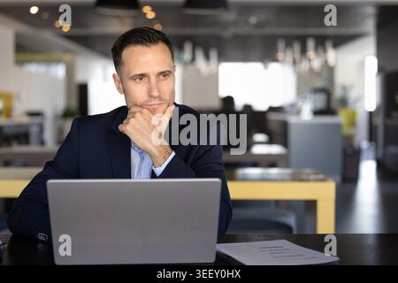 Businessman sitting at desk working on laptop with documents nearby Stock Photo