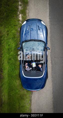 Aerial view of a blue convertible car parked on a roadside with three people looking up from inside next to a green grassy field La Chapelle-de-Guinch Stock Photo