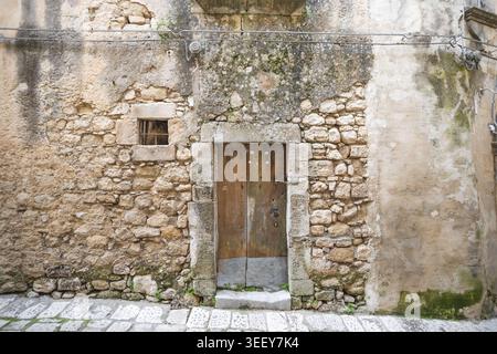 View of a rustic wooden door set in an ancient stone wall with a small barred window and mossy textures in Ragusa, Sicily, Italy. Stock Photo