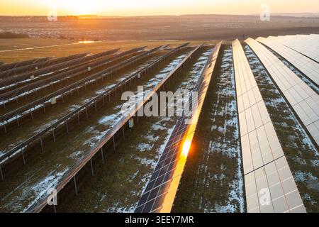 Photovoltaic solar panels aligned systematically on grassy field during early morning light. Snow patches visible on ground with soft sunlight reflecting off panel surfaces. Stock Photo