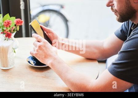 Man using smartphone while sitting at cafe table with cup of coffee, close up view, focused on screen, cozy interior with flowers, natural light, concept of online communication and modern lifestyle Stock Photo