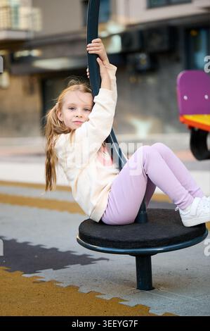 Girl sitting on playground spinner holding pole, smiling at camera, active outdoor play, urban background, casual outfit, childhood fun Stock Photo