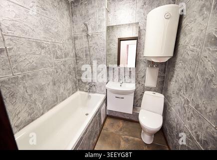 Modern bathroom interior with bathtub, toilet and compact sink vanity. Small washroom with gray textured walls, mirror, wall mounted water heater and ceramic floor Stock Photo