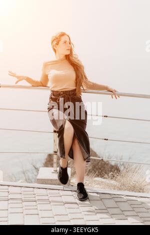 Woman ginger coastline leaning on a metal railing at a sea viewpoint during a bright sunset enjoying the outdoor scenery Stock Photo
