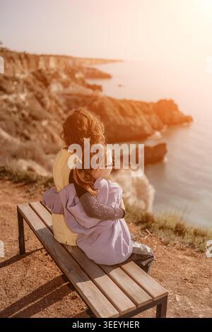 Mother child sunset woman and girl sit on a bench looking at the sea from a scenic cliff edge during golden hour travel Stock Photo