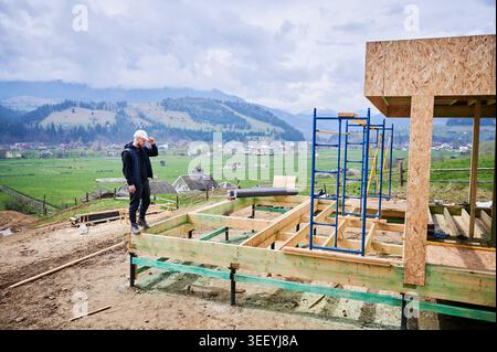 Construction worker standing on wooden foundation and surveying rural building site, concept of project planning, site evaluation, development progress and countryside housing Stock Photo