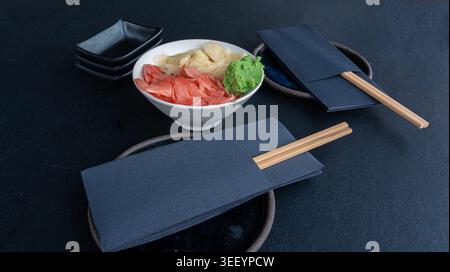 Pickled pink and white ginger and wasabi bowl on dark table, traditional Japanese condiments served with sushi, stacked sauce dishes and napkins on bl Stock Photo