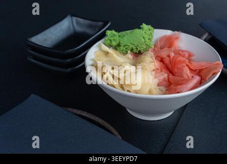Pickled pink and white ginger and wasabi bowl on dark table, traditional Japanese condiments served with sushi, stacked sauce dishes and napkins on bl Stock Photo