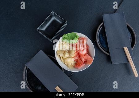 Pickled pink and white ginger and wasabi bowl on dark table, traditional Japanese condiments served with sushi, stacked sauce dishes and napkins on bl Stock Photo