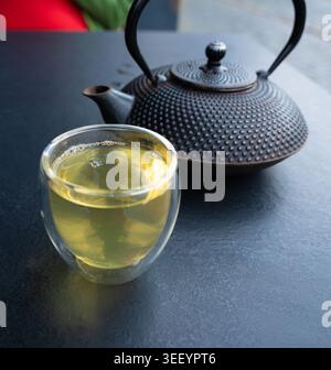 Glass cup with green tea and cast iron teapot on dark table. Traditional Asian drink served in modern double wall glass, tea time setting for menu, ca Stock Photo