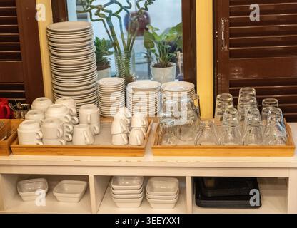 Hotel dining area, stacked plates, cups, glasses, trays, organized setup, breakfast service, white ceramic, glassware, self-service station kitchenwar Stock Photo