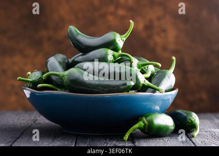 Jalapeno peppers piled in blue bowl on wooden surface with warm brown backdrop. Fresh chili, organic vegetables, rustic composition, food still life Stock Photo