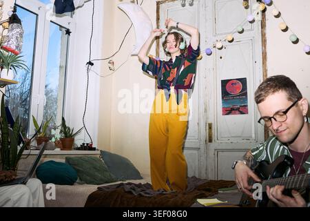 Young adult woman dancing in shared apartment while young adult man playing guitar nearby, expressing creative leisure and friendship. Useful for youth lifestyle or home entertainment marketing Stock Photo