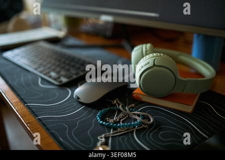 Close up of desktop workspace showing headphones, keyboard, mouse, notebook, jewelry on desk mat, suggesting remote work routine. Supporting productivity, content creation, lifestyle branding Stock Photo
