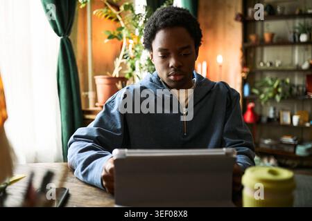 Medium shot of Black teenager using tablet at home, studying online and focusing on digital learning. Suitable for education technology, remote classes, youth learning campaigns Stock Photo