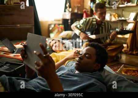 Young Black man relaxing with tablet while friends resting and playing guitar in shared apartment, representing digital leisure and youth lifestyle. Ideal for social media or streaming ads Stock Photo