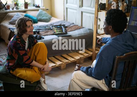 Young adult Caucasian woman and Black man discussing coding project in home workspace, representing collaboration and tech learning. Useful for remote work, startup, mentoring campaigns, flat share Stock Photo