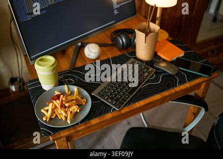 Close up of home office desk showing coding monitor, keyboard, smartphone, coffee cup, fries and headphones, illustrating remote work routine. Useful for productivity or work life balance Stock Photo
