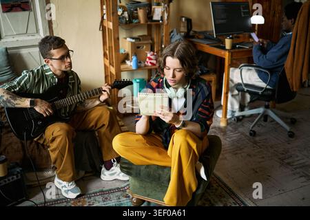 Young adults collaborating at home, woman writing song lyrics while man playing electric guitar and roommate working on computer. Ideal for creative teamwork, remote lifestyle marketing Stock Photo