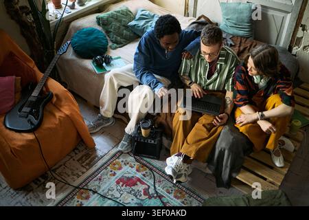 High angle view of diverse young adults collaborating on laptop in shared apartment, discussing creative project and teamwork. Ideal for remote work, startup, digital collaboration Stock Photo