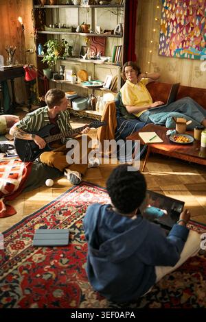 Young adults collaborating in living room, playing guitar and using laptop and tablet, showing creative teamwork and casual content production. Useful for remote collaboration or creator economy Stock Photo