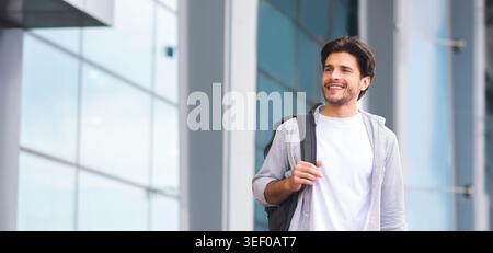 Smiling Young Man With Backpack Standing Outdoors Near Airport Building Stock Photo