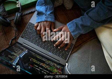 Close up of Black young adult typing on laptop with coding screen and headphones, working remotely on software project. Ideal for remote work, programming, tech training marketing Stock Photo