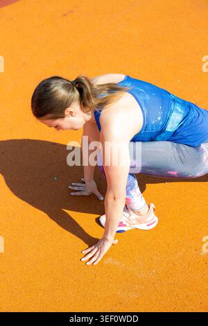 High angle view of a fit woman in colorful leggings performing deep low lunge stretch on outdoor sports ground. Professional athletic warm-up routine. Stock Photo