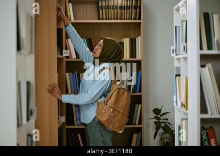 Adult Muslim Woman Reaching for Book in Library for Education Stock Photo