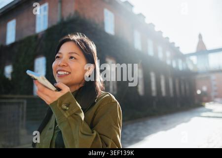 Smiling young Asian woman commuting through city streets, speaking into her phone to record a voice message while walking past historic brick building Stock Photo